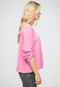 Maglietta rosa a maniche lunghe con maniche arrotolate e dettaglio dei bottoni, vestibilità ampia e texture morbida. Indossata con jeans grigio scuro.