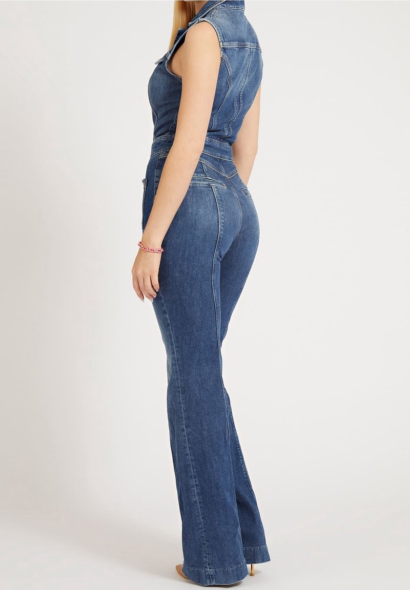 Femme portant une combinaison en jean sans manches avec une taille cintrée et des jambes évasées, debout de profil sur un fond uni.