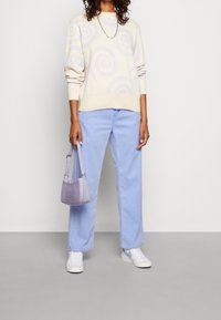 Cremefarbener Pullover mit lila Wirbelmuster, hellblaue Jeans und eine kleine lila Tasche. Weiße Sneaker mit strukturierter Oberfläche.