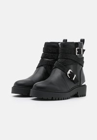 Botas de cuero negro hasta el tobillo con correas texturizadas negras y herrajes de plata. Presentan una suela de goma resistente y una cremallera lateral para facilitar su uso.