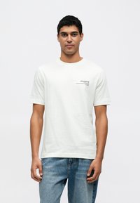 Biała bawełniana koszulka z krótkimi rękawami, z małym czarnym logo na lewej piersi i okrągłym dekoltem. Nosi się ją z niebieskimi dżinsami.