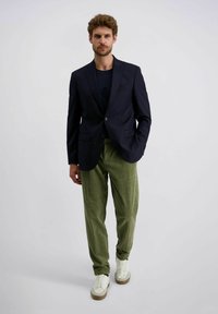 Man loopt in een marineblauwe blazer, zwart shirt, olijfgroene corduroy broek en witte sneakers tegen een effen grijze achtergrond.