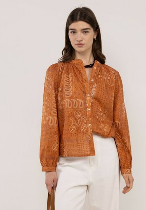 Mujer con blusa abotonada de manga larga con estampado de cachemira naranja y pantalones blancos, sosteniendo un bolso marrón a su lado.