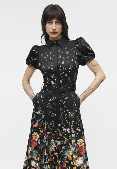 Femme aux cheveux foncés portant une robe midi noire à manches bouffantes avec un imprimé floral et papillons coloré, les mains dans les poches, fond neutre.