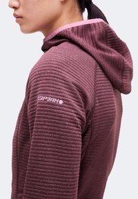 Personne portant une veste à capuche côtelée bordeaux avec une doublure rose à l'intérieur de la capuche et un petit logo sur la partie supérieure de la manche.
