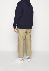 Námořnicky modrá mikina s kapucí, béžové ohrnuté kalhoty, bílé boty se zelenými akcenty. Materiál působí měkce, s volnějším střihem. Jednoduchý design.