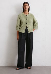 Hellgrüner, flauschiger Strickcardigan mit rundem Ausschnitt und fünf dunklen Knöpfen. Getragen über lockeren schwarzen Hosen und Slidesandalen.