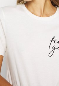 Bílý krátký rukáv tričko s kulatým výstřihem a černým kurzívou napsaným "feel good" na přední straně. Měkká textilie.