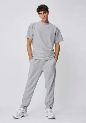 Jeune homme debout les mains dans les poches, portant une chemise à manches courtes gris clair, un pantalon assorti et des baskets blanches New Balance.
