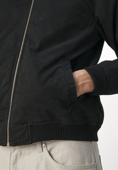 Chaqueta bomber negra con cremallera frontal, puños y dobladillo acanalados. La tela parece suave con un acabado mate. Cuenta con un bolsillo lateral.