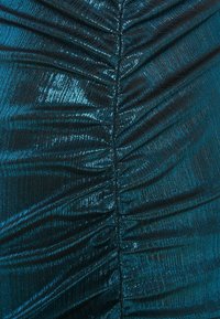 Close-up van glanzende stof in teal en zwart met een verzamelde naad die verticaal door het midden loopt, waardoor textuurvouwen en hoogtepunten ontstaan.