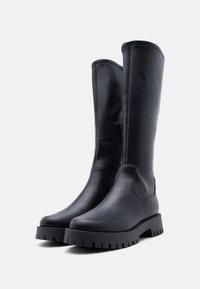 Ζεύγος ψηλών μαύρων δερμάτινων μποτών με χοντρά πέλματα και πλευρικά φερμουάρ, όρθιες σε απλό φωτεινό φόντο.