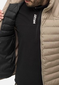 Beige gepolsterte Jacke mit steppdesign, schwarzem Innenfutter und Reißverschluss. Über einem schwarzen Millet-Fleece-Oberteil getragen.