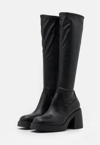 Musta polven korkeudelle ulottuva saappaat, jotka on valmistettu sileästä nahasta. Varustettu paksulla korolla ja sivuvetoketjulla. Kiiltävä pinta ja tiukka muotoilu.