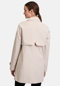 Beżowy płaszcz trench z tylnym detalem, wykonany z gładkiej tkaniny, z długimi rękawami i luźnym krojem. Minimalistyczny design bez wzorów.
