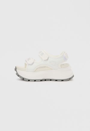 Sandalia blanca de plataforma con suela gruesa gris con dibujo y tiras ajustables de velcro en la zona de los dedos y el tobillo.