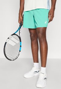 Αθλητικά σορτς σε μέντα πράσινο, ύφασμα που απομακρύνει την υγρασία, ελαστική μέση, συνδυασμένα με μαύρη και μπλε ρακέτα τένις και λευκά αθλητικά παπούτσια.