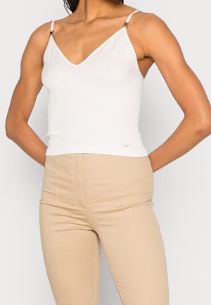 Kobieta ubrana w biały, bezrękawnik z regulowanymi ramiączkami oraz beżowe, dopasowane spodnie z wysokim stanem na jednolitym tle.
