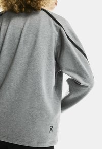 Person with curly hair wearing a loose grey sweatshirt with black stripe on shoulder and logo near hem, facing away with hand on hip.