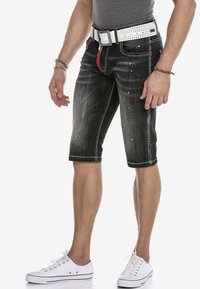 Schwarze Denim-Shorts mit verblassten Bereichen, kurzer Schnitt, weißen Nähten und roten Akzenten an der Tasche; kombiniert mit einem weißen Nieten-Gürtel und Sneakers.