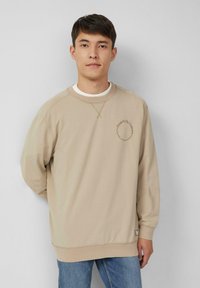Beige sweatshirt met een ronde halslijn, ribgebreide manchetten en zoom. Bevat gestikte details en een geprinte afbeelding op de borst. Gedragen met blauwe jeans.