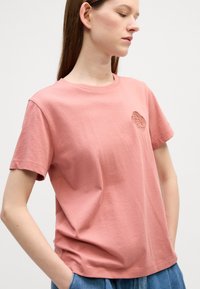 Kortskaftet rosa bomull t-skjorte med rund hals og en brodert abstrakt design på venstre bryst, med en avslappet passform.