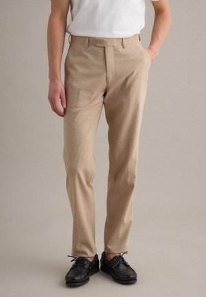 Hombre con pantalones beige con cierre de botón y cremallera, combinados con zapatos negros de cordones y una camisa blanca metida dentro.