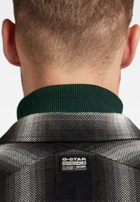 Giacca houndstooth nera e grigia con colletto a coste verde; presenta un'etichetta G-Star Originals sul retro.