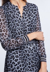 Kobieta w długim rękawie, zapinanej sukience w panterkę z prążkowanymi mankietami, opierająca dłoń na przeciwnym ramieniu na jednolitym tle.