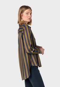 Blusa a righe in blu scuro e giallo senape, con colletto a bottoni, maniche lunghe e orlo posteriore leggermente più lungo.