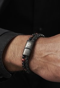 Schwarzes geflochtenes Lederarmband mit schwarzen und roten Perlen, ausgestattet mit einem metallischen Verschluss, der ein Logo-Detail aufweist.