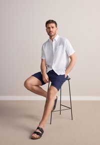 Chemise blanche en lin à manches courtes avec une poche, associée à un short bleu marine et des sandales noires. Homme assis sur un tabouret en métal noir.