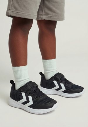 Kind draagt zwart-witte sportschoenen met witte sokken en beige korte broek tegen een effen achtergrond.