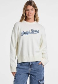 Pulover alb din tricot cu tiv și mansete cu dantelă; prezintă logo-ul brodat "Guess Jeans" în culoare albastră pe piept; are un croi lejer și un guler rotund.