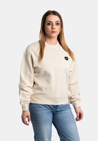 Crème-kleurige sweatshirt met een ronde hals, ribgebreide manchetten en zoom. Heeft een klein zwart hartlogo op de borst. Gekocht met lichtblauwe jeans.
