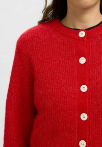 Röd cardigan i mjukt stickat material med rundad halsringning, knappframstängning med fyra vita knappar och textil med struktur.