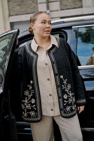 Femme avec les cheveux en chignon sortant d'une voiture noire portant une tenue beige boutonnée et une veste matelassée noire brodée de fleurs blanches.