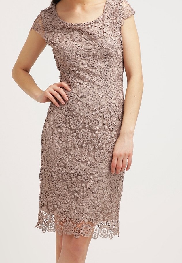 Femme portant une robe en dentelle mauve longueur genoux avec des motifs floraux circulaires et des manches courtes, debout avec une main sur la hanche.
