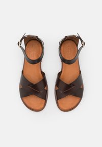 Braune Ledersandalen mit einem gekreuzten Riemendesign, offenen Zehen und einem Knöchelriemen mit einer Messingschnalle. Beigefarbene Fußsohle mit strukturierten Details.