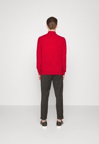 Pull en laine rouge à col montant, associé à un pantalon foncé et des baskets. La coupe est décontractée et la texture semble douce.
