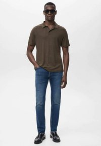 Polo de manga corta marrón combinado con jeans azules. Modelo lleva gafas de sol negras y zapatos brillantes negros. Diseño simple y ajustado.
