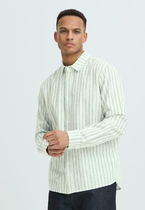 Homme portant une chemise à manches longues rayée de couleur claire, boutonnée, avec un pantalon foncé, se tenant devant un fond uni.