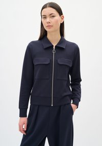 Giacca zip con colletto di colore navy, due tasche sul petto e cintura aderente. Tessuto liscio e accessori in argento accentuano il design.