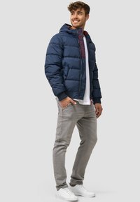 Chaqueta acolchada navy con capucha, textura de amortiguación, marca en rojo en el zipper, combinada con jeans gris claro y zapatillas blancas.
