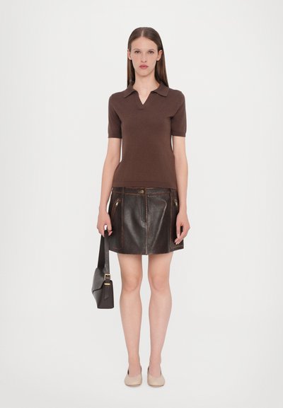 Polo shirt à manches courtes marron associé à une mini-jupe en cuir marron avec des poches zippées à l'avant. Le mannequin tient un sac à bandoulière noir.