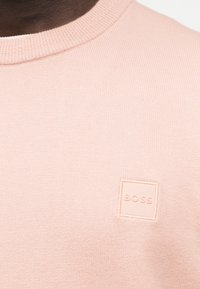 Κοντινή λήψη ανοιχτό ροζ πλεκτού πουλόβερ με στρογγυλή λαιμόκοψη και μικρό τετράγωνο λογότυπο BOSS στο στήθος.