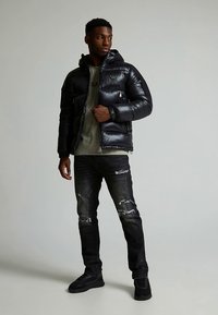 Juoda pūkinė striukė su kapišonu, kišenėmis su užtrauktukais ir logotipais; derinama su suplėšytais tamsiais džinsais ir juodais sportbačiais. Lygi, blizgi tekstūra.