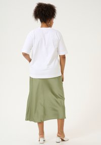 Un haut blanc à manches courtes associé à une jupe satin olive lâche, avec une texture lisse. La tenue est complétée par des sandales blanches à talons.