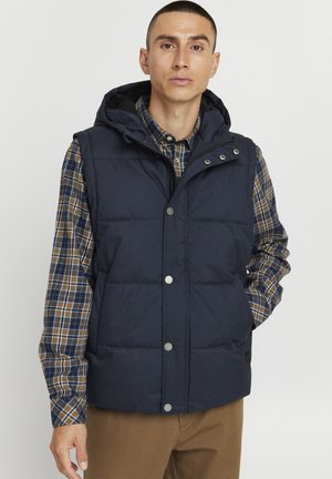 Gilet rembourré marine avec capuche, doté de boutons-pression, d'un design matelassé et d'un tissu résistant. Porté sur une chemise à carreaux à boutons.
