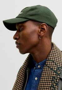 Green baseball cap with a curved brim, worn with a checkered scarf and denim shirt. Features a hoop earring on the left ear.